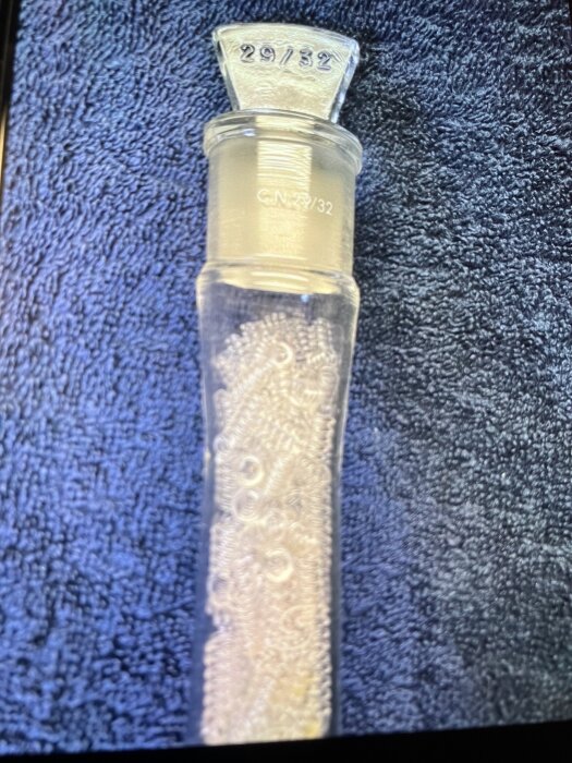 Glasflaska med spiralformad insats och markeringarna "29/32" och "CN 29/32", placerad på blå textilbakgrund.