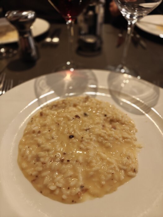 Krämig risotto serverad på en vit tallrik, omgiven av vinglas och restaurangbestick.