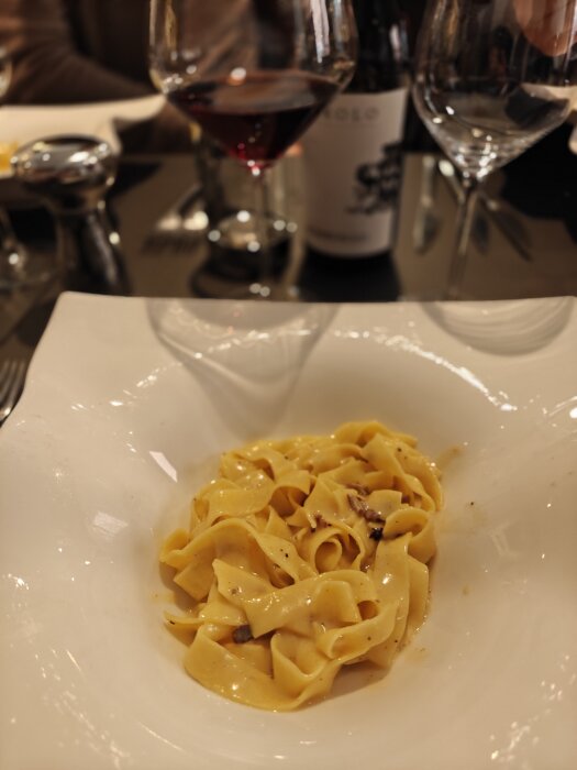 Pasta med tryffel och vit sås i djup tallrik, omgiven av vinglas och vinflaska på middagsbord.