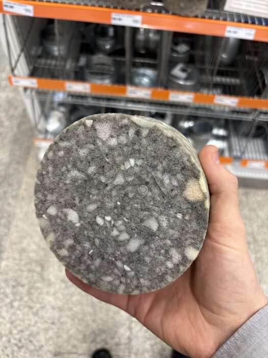 Hand håller en rund, grå skumgummikudde i en järnaffär, framför ett ställ med hyllor i bakgrunden.