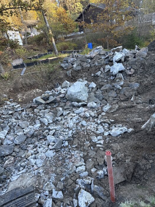 Hög av stenar och grus på byggarbetsplats med träd och hus i bakgrunden, röd avgränsningsstång synlig i förgrunden.