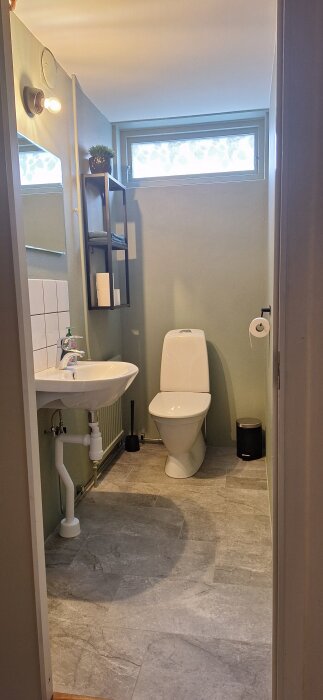 Nyrenoverad källartoalett med grå klinkergolv, ny färg på väggar, handfat, toalettstol, och hylla bredvid fönster.