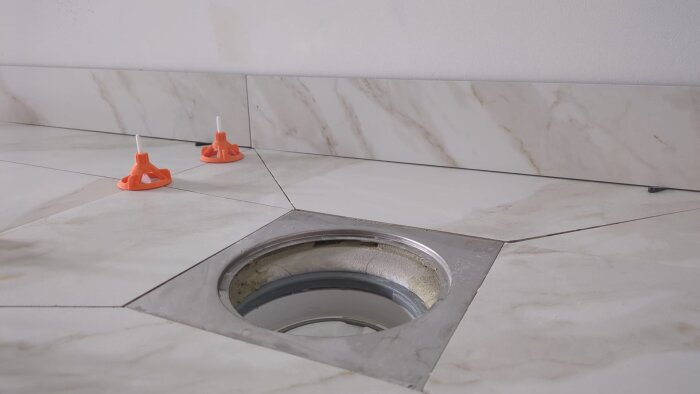 Kakelplattor lagda runt ett golvbrunnsområde i en tvättstuga, med synligt tätskikt och två orange distansclips på plattorna.