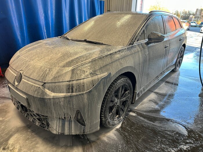 En bil täckt i skum under tvätt i en tvätthall, med ett blått draperi i bakgrunden.