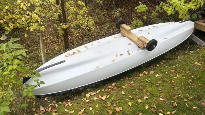 Vit trampbåt uppochner i trädgård med rullvagn av trä och hjul, omgiven av höstlöv och grönska.