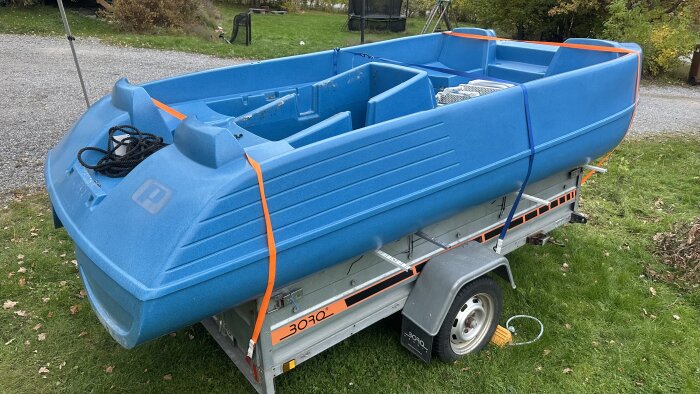 Blå trampbåt, Pioner Trio, fastspänd på en släpvagn, placerad på gräsmatta nära grusväg.