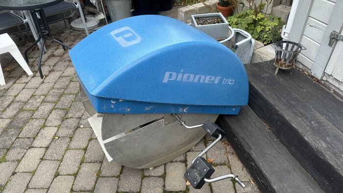 Blå trampbåt Pioner Trio uppställd på en stenlagd yta med växter och trädgårdsmöbler i bakgrunden.