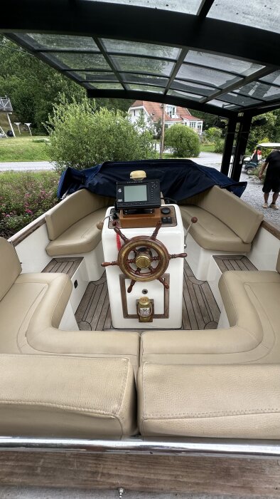 Båtinteriör med roder och ratt, omgiven av beige sittplatser under ett genomskinligt tak, samt en skärm monterad på konsolen.