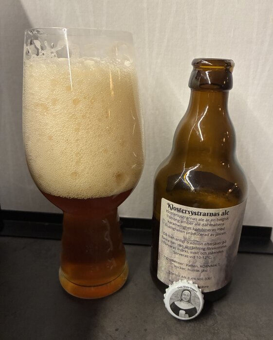 Ett glas fyllt med skummande öl bredvid en brun ölflaska med etikett som beskriver ölens egenskaper, samt en kapsyl med nunna.