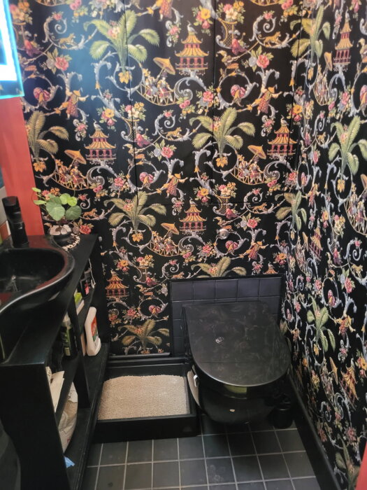 Toalett med svart och färgglad mönstrad tapet, svart klinkergolv och dold förvaring ovanför toaletten.