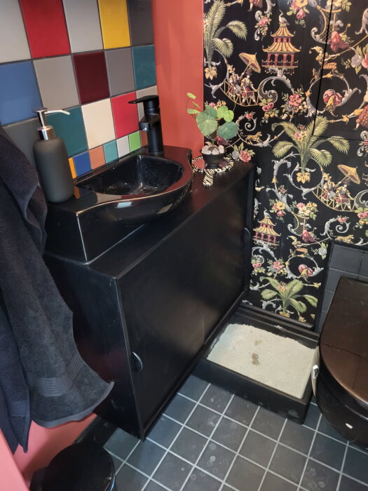 Svart badrumsmöbel med tvättställ, färgglatt kakel och blommig tapet.