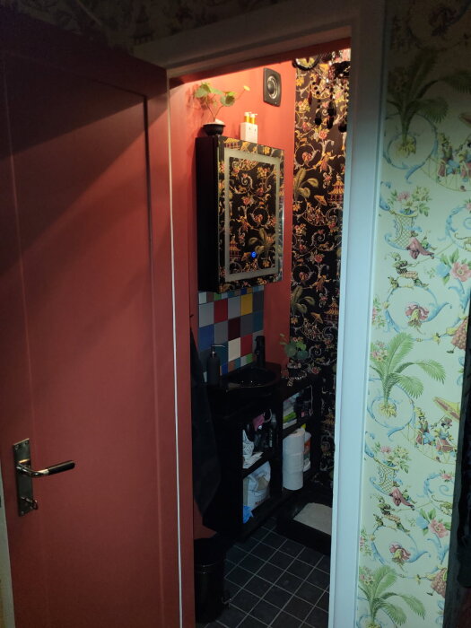 Badrum med mönstrade tapeter, klinkergolv, och förvaring ovanför toaletten. Svart handfat med spegel.