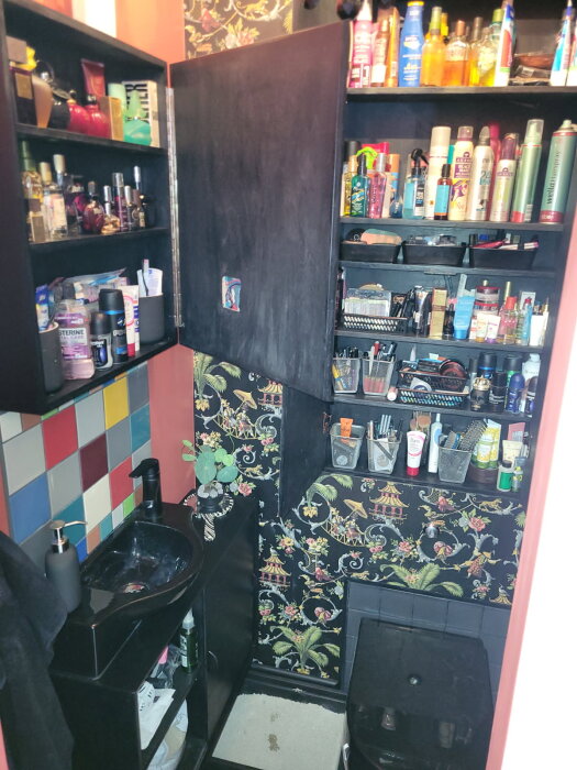 Liten toalett med svart inredning, färgglada kakelplattor och blommig tapet. Öppen skåpvisning med dolda hyllor ovanför toalettstolen.