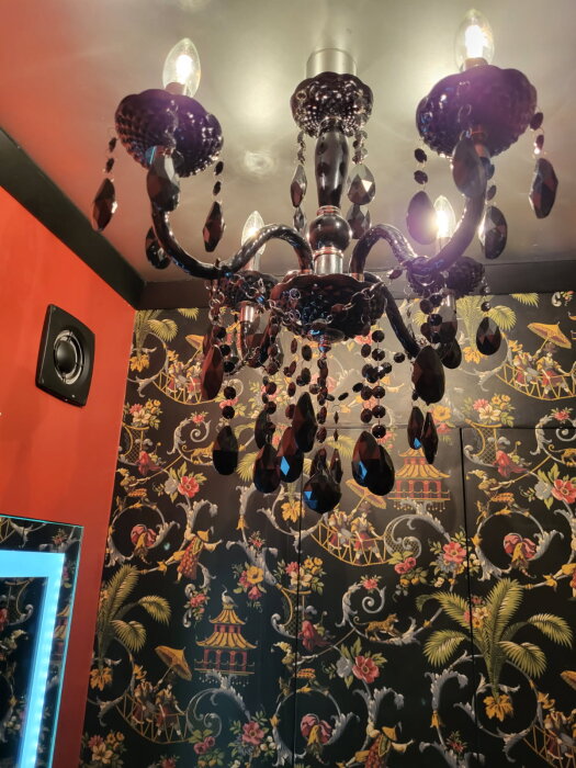 Svart kristallkrona i ett badrum med mönstrad tapet och röd vägg, spegel med inbyggd belysning syns delvis.