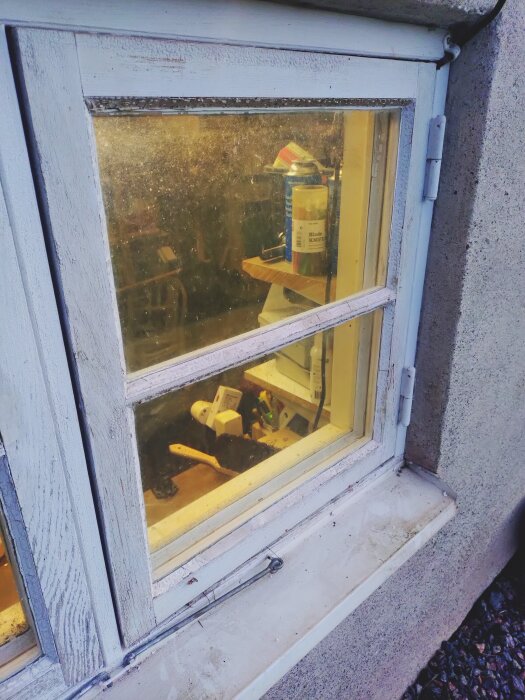 Fönster med trasigt glas i en blåmålad ram, sett från utsidan. Verktyg syns på hylla genom fönstret.