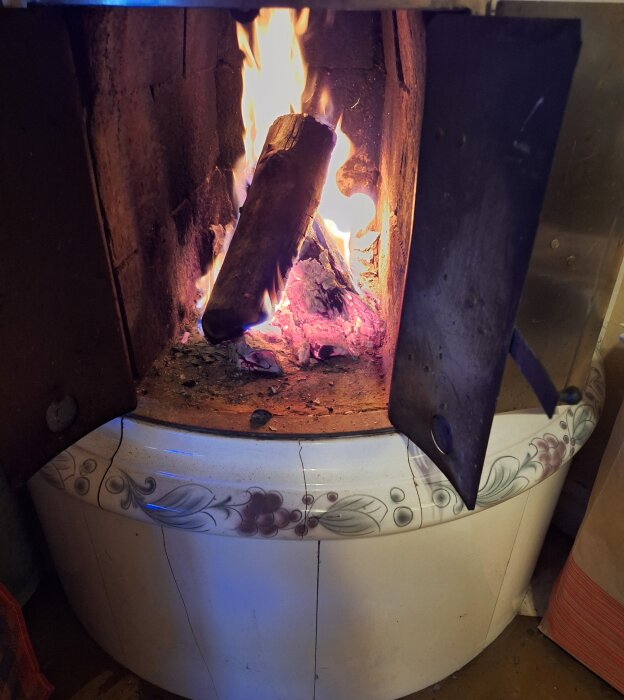 Eld i en vit kakelugn med blommönster, värmande vedträn som brinner för höstens första brasa.