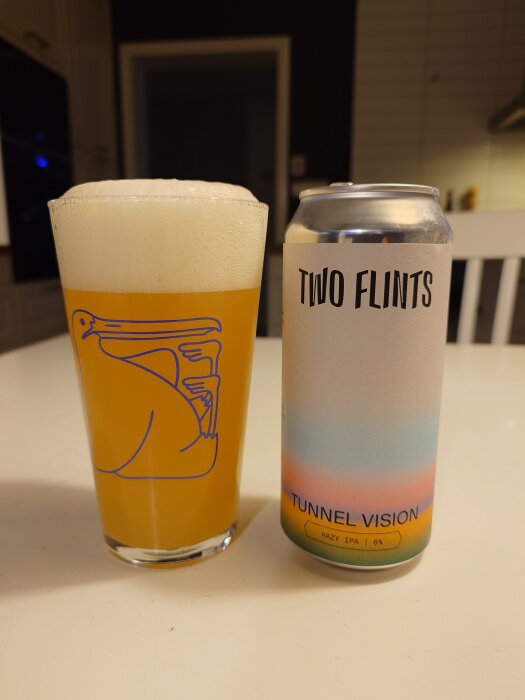 Ölglas med skummande öl bredvid en burk Tunnel Vision Hazy IPA från Two Flints på ett bord.