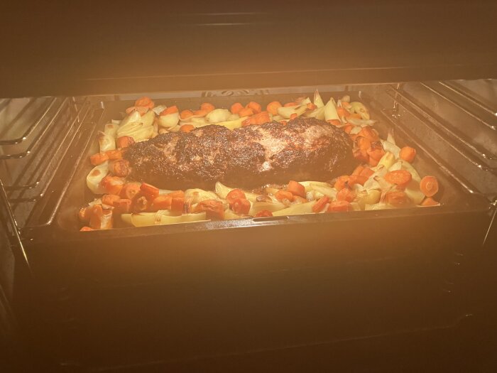 Köttfärslimpa i ugnen omgiven av ugnsbakade potatisar och morötter.