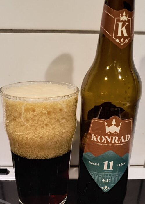 Mörk öl med skummande spets i ett glas bredvid en flaska med etiketten "Konrad Tmavý Ležák 11".