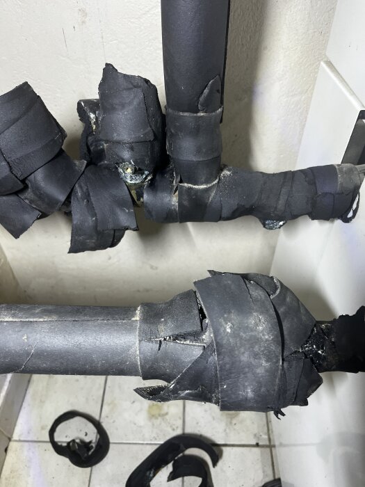 Svarta rör isolerade med tejp, delvis borttagna, exponerar kanske köldmedieslang eller anslutning nära kakelklädd vägg.