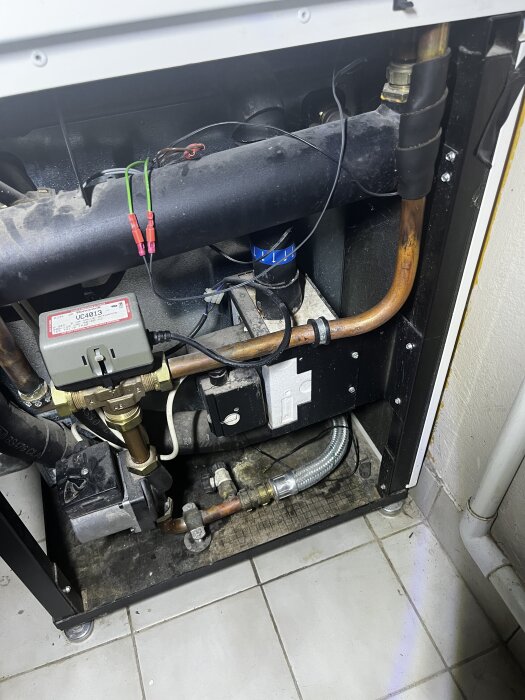 Inre delen av en värmepump med rör och kopplingar, synlig isoleringstejp, och en magnetisk slang längst ner i en verkstadsmiljö.