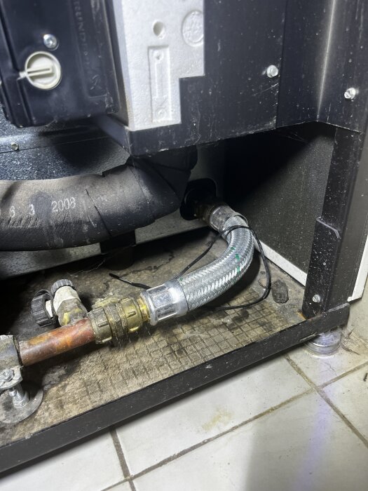 Grå slang och ventil vid pumpens botten, delvis isolerad och ansluten till en kopparrör, på ett kakelgolv.