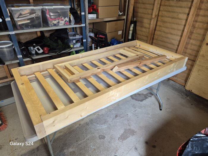 Ram av trä för en barnvänlig säng under byggnad i ett garage, placerad på ett hopfällbart bord med verktyg och material i bakgrunden.