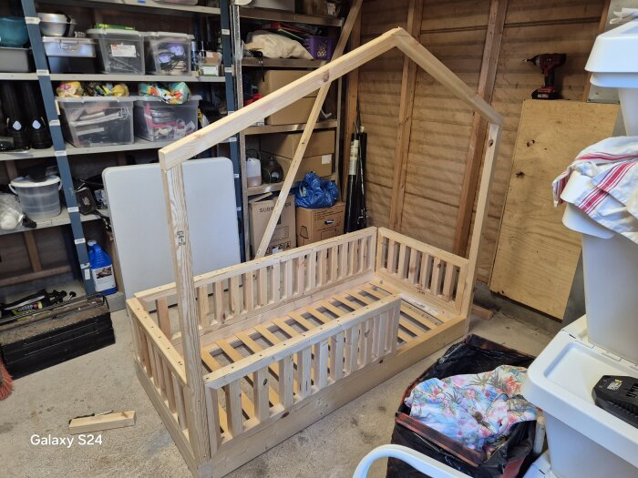Träram för en barnsäng under konstruktion i ett verkstadsutrymme med verktyg och förvaringshyllor i bakgrunden.