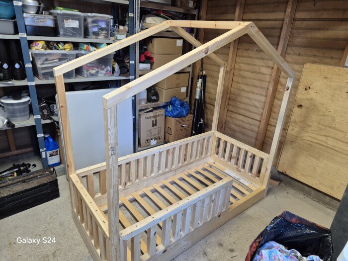 Träram för en barnsäng med husdesign under uppbyggnad i ett garage, omgiven av verktyg och förvaringshyllor.