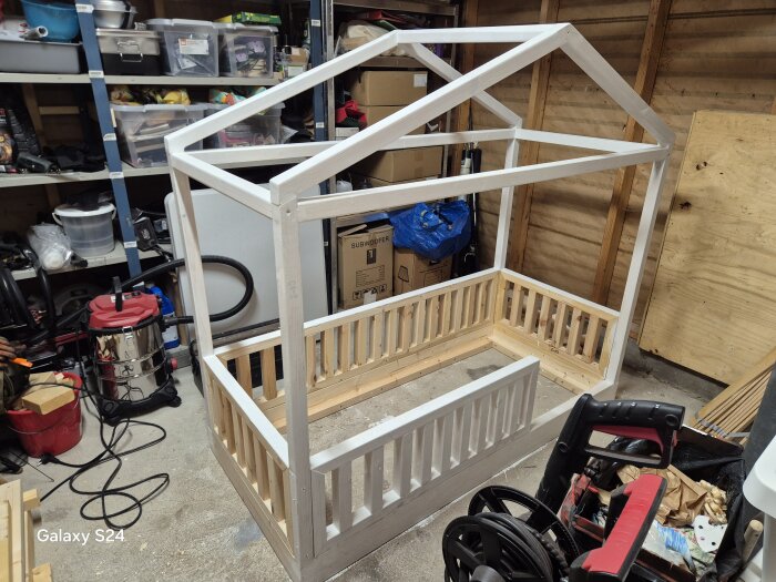 Hemmagjord barnsängsram med hustak i trä, under byggande i en verkstad fylld med verktyg och material.