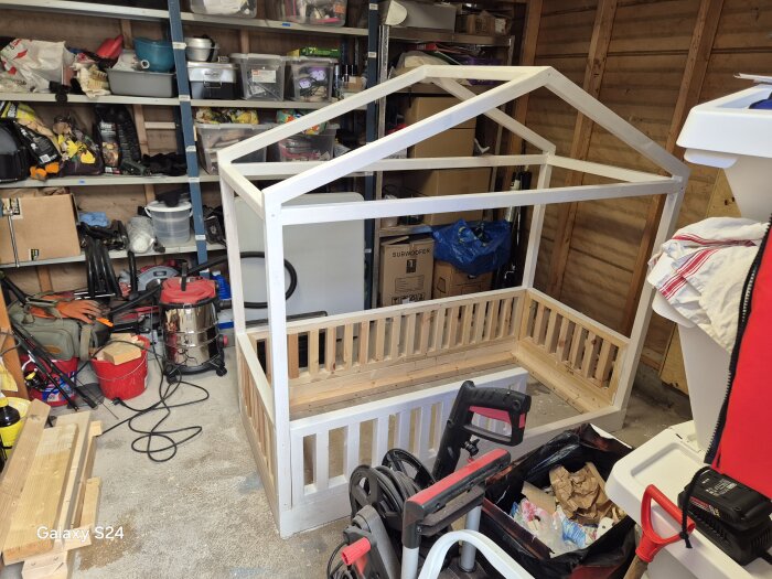 Barnsäng med husformad ram byggs i garage, omgiven av verktyg och byggmaterial.