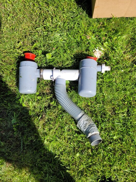 Grått vattenlås med röd plastmutter och slang på gräsmatta, del av diskussion om byte av äldre vattenlåssystem till modernare lösning.