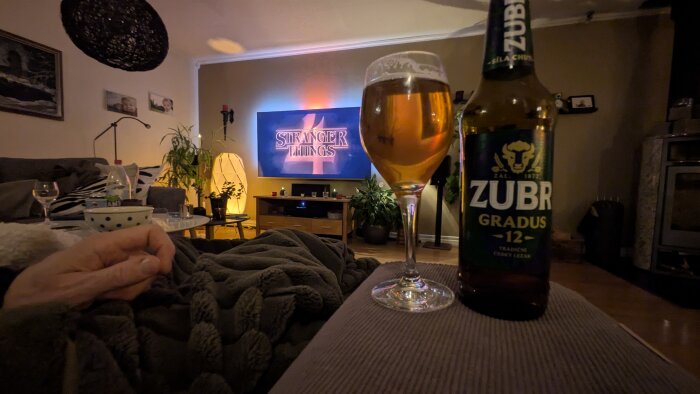 Flaska och glas med Zubr-öl framför en TV som visar Stranger Things säsong 4, i ett mysigt rum med inredning och växter.