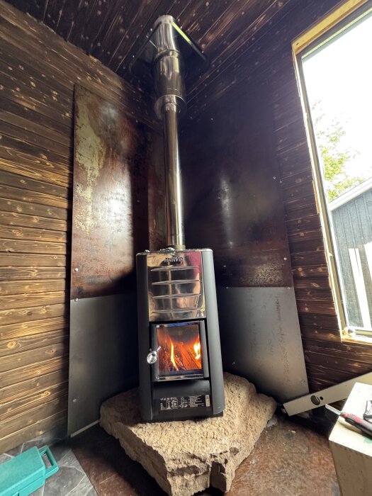 Kamin placerad på stor ölandsten med brinnande eld i, omgiven av mörkt träpanel och metallplåtar.