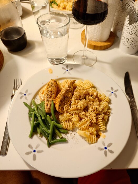 Tallrik med dragonkyckling, pasta och haricots verts på ett bord. Glas med vatten och rödvin bredvid tallriken.