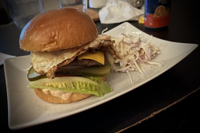 Hamburgare med dressing, gurka, ost, sallad, tomat, stekt och rå lök, senap och ägg serverad med coleslaw på en tallrik.