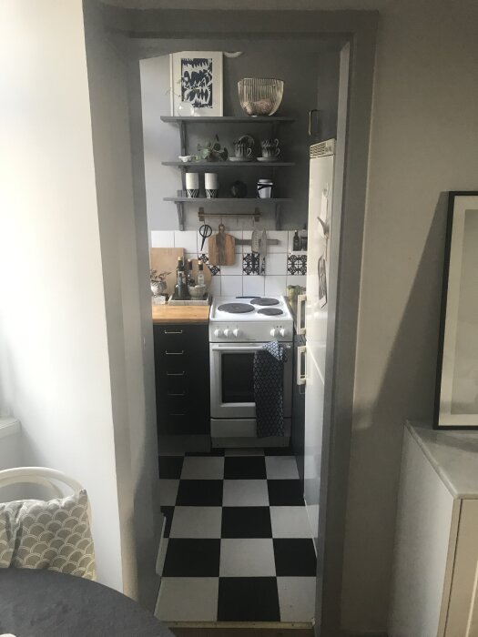 Litet kök med svartvita rutiga golvplattor, öppen hylla med koppar och skål, spis med blå handduk och trähylla med köksredskap.