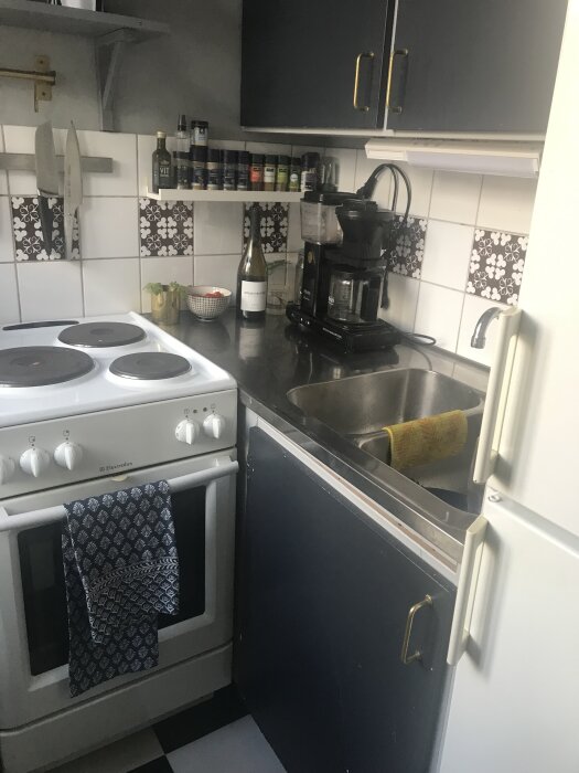 Litet kök med vit spis, svart kaffekokare, mörka skåpluckor, hylla med kryddor och vit kakelvägg med svartvit mönstrad dekor.