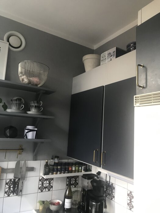 Grått kök med svarta skåpluckor, hylla med porslin och kryddhylla, kaffemaskin på bänkskiva, vit väggventil och pastamaskinslåda ovanpå skåpen.