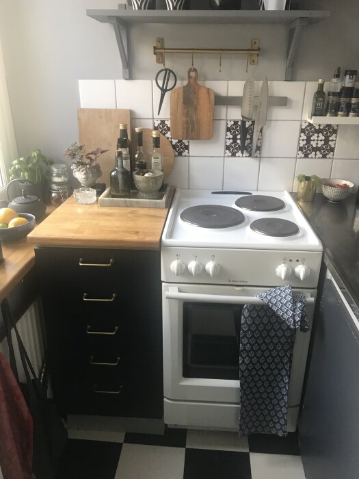 Litet kök med spis, svart-vita golvplattor, mörka skåp, träskivor och hyllor. Köksredskap och kryddor synliga.