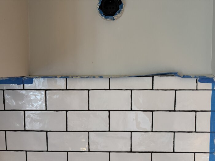 Vitt kakel på en köksvägg, maskeringstejp längs kanten och en öppning för fläktinstallation. Problem med kakel som sticker ut.