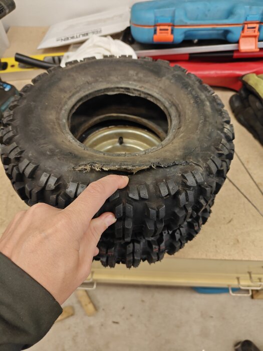 Däck till snöslunga med slitage i fokus, hand pekar på skada. Verktyg och verkstadsmaterial syns i bakgrunden.