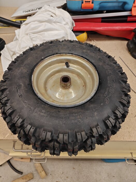 Däck med fälg för en reservsnöslunga, placerad på en arbetsbänk omgiven av verktyg och material.