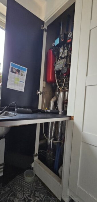 Fjärrvärmesystem monterat i en vit skåp, exponerat med dörren öppen, synligt rörsystem, golv med mönstrade klinkers, del av diskbänk och vägg.