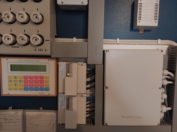 Kontrollpanel med knappar och en display märkt "Husdator EMD 4120" monterad på blå vägg tillsammans med annan elektrisk utrustning.