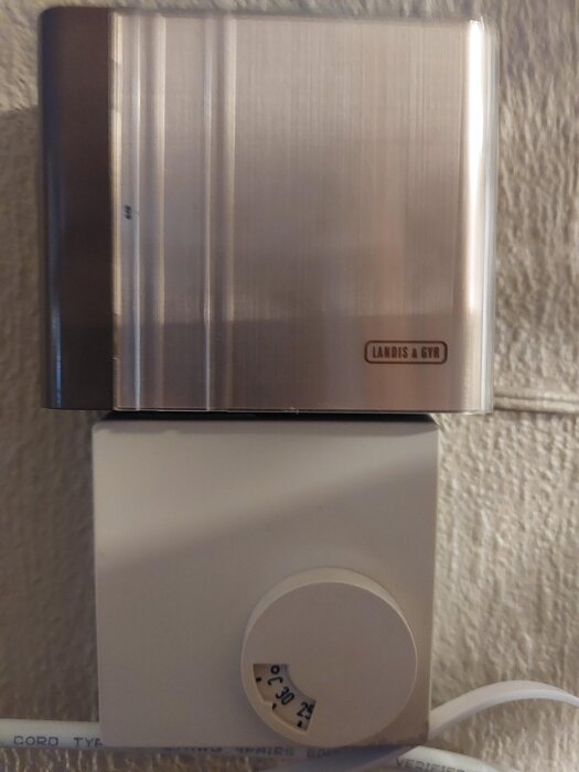 Väggmonterad termostat av märket Landis & Gyr med en ratt som visar en temperaturinställning mellan 25 och 30 grader Celsius.