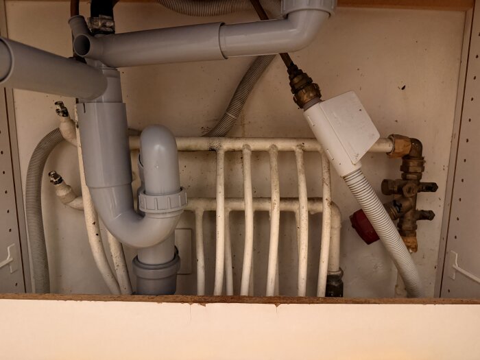 Under diskbänk visas grå avloppsrör och vita värmerör, del av ett värmesystem i en lägenhet, med ventiler och kopplingar synliga.