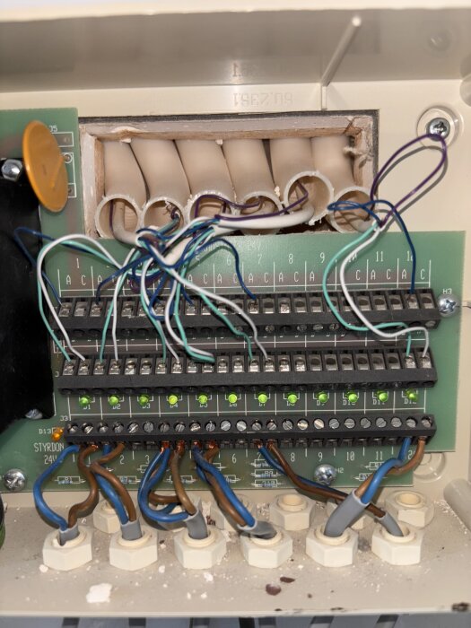 Anslutningar och kablar i en styrbox för golvvärme. Flera gröna lampor är tända, och kablarna är anslutna till plintarna.