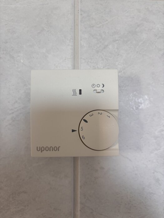 Termostat från Uponor för golvvärme, monterad på en kaklad vägg med en vridbar ratt för temperaturinställning och symboler för olika lägen.
