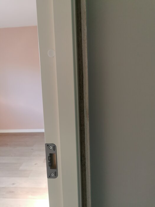 Närbild på en dörrkarm med synlig glipa mellan karmen och väggen, i ett rum med trägolv och ljusa väggar.